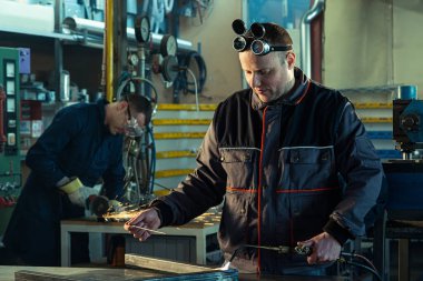 İş arkadaşı arka taraftaki öğütücüde fark edilirken kaynakçı atölyede otomatik kaynak aleti ile çalışıyor..