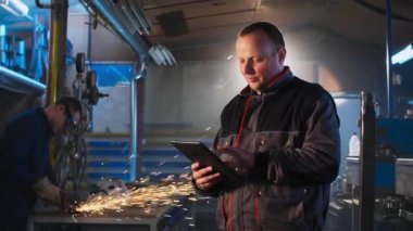 Atölyesinde tableti olan tamirci bir şeyler yazarken, arka planda meslektaşı öğütücü üzerinde çalışıyor..