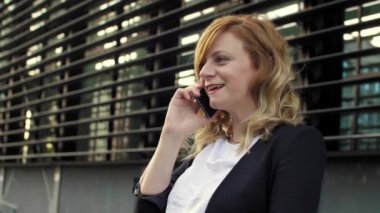 Modern bir binanın önünde cep telefonuyla konuşurken gülümseyen hamile bir iş kadını. Kol saatine bakıyor..