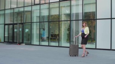 Modern bir binanın önünde elinde bir parça seyahat valiziyle yürüyen hamile, beyaz bir kadın kol saatine bakıyor..