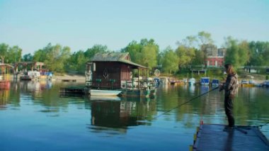 Uzun saçlı bir adam salla nehir kenarında balık tutuyor. Etrafında kayıklar ve yüzen evler ve arka planda ağaçlar var. Balıkçı oltası kullanıyor, makaralı, yeşil ipli ve yemli..