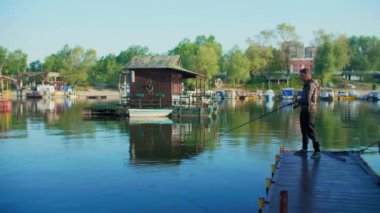 Uzun saçlı bir adam salla nehir kenarında balık tutuyor. Etrafında kayıklar ve yüzen evler ve arka planda ağaçlar var. Balıkçı oltası kullanıyor, makaralı, yeşil ipli ve yemli..