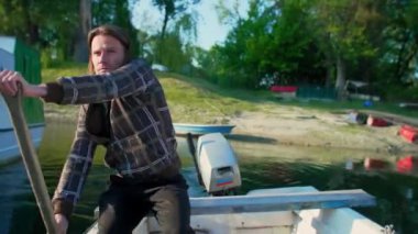 Beyaz orta yaşlı adam eski beyaz teknesinde kürek çekiyor. Gün doğumunda nehir marinasında. Bu balıkçı eski tahta küreği kullanıyor. Etrafında kayıklar ve yüzen evler ve arka planda ağaçlar var..