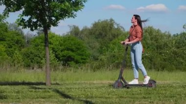 Genç ve güzel bir kadın parkın içinde elektrikli scooterla geziyor ve yeşil bir çevrede, bazı çekimlerde saçlarında rüzgar var..