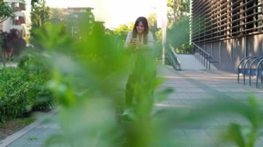 Telefonunu kullanırken elektrikli scooter 'ın yanında duran genç ve güzel bir kadın. Gülümsüyor, iş elbisesi giyiyor, arkasında modern mimari figürler var..