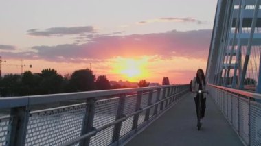 Genç ve güzel bir kadın elektrikli scooterıyla köprüden geçiyor. İş eğitimi almış ve gülümsüyor. Arkaplan, modern bir köprünün parçalarından oluşuyor..