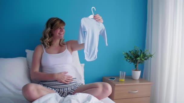 小さな赤ん坊の服を見ながら彼女のベッドの中で座っている位置に笑顔と幸せな妊娠中の白人女性 — ストック動画