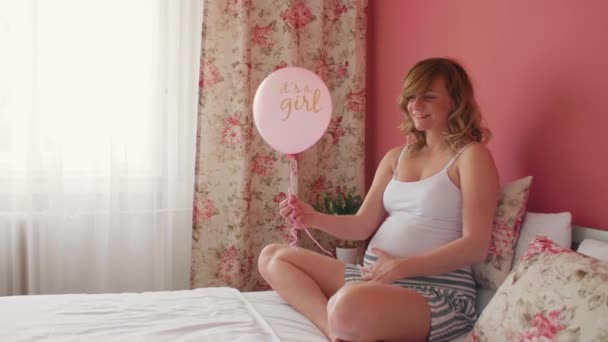 一个快乐的孕妇抱着气球躺在床上 上面写着 是个女孩 她一只手放在肚子上 — 图库视频影像