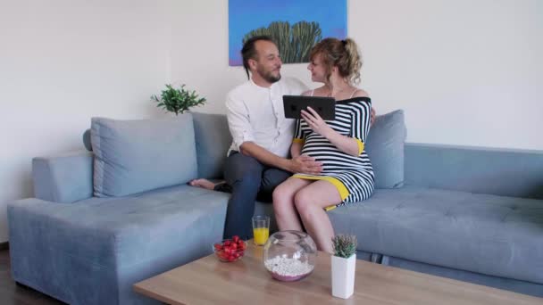 一对年轻貌美的已婚怀孕夫妇正坐在客厅的沙发上看平板电脑 — 图库视频影像