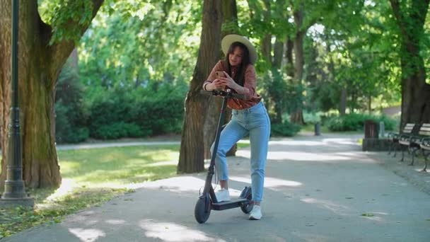一位年轻美丽的女人 戴着一顶帽子 站在她的电动车旁边 一边在手机上打字 背景以树木和绿色为主 — 图库视频影像
