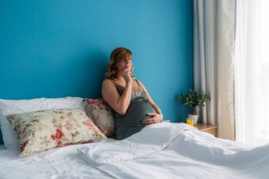 Yarı-oturma pozisyonunda, elleri karnına dolanmış, bebeği merak eden hamile bir kadın. Çok düşünceli görünüyor..