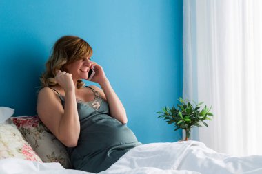 Akıllı telefonuyla konuşurken yarı oturduğu bir pozisyonda hamile bir kadın. O, aldığı haber hakkında mutlu ve heyecanlı görünüyor..