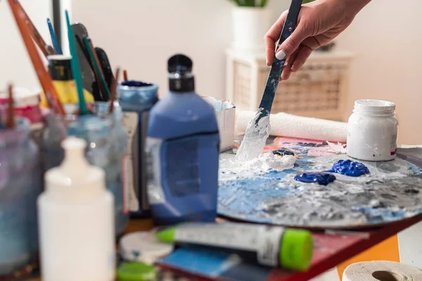 The painter\'s hand smears paint on a palette in her home studio. The paint pots and the painting brush are in jars on the side.