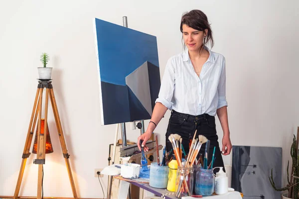 In her studio, a young smiling painter smears paint with a paintbrush on a palette. Around it is a work of art placed on an easel, as well as other accompanying material for painting.
