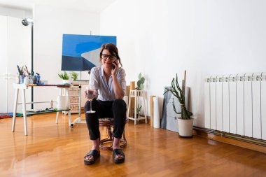 Genç bir bayan ressam ev stüdyosunda oturuyor ve cep telefonuyla konuşuyor. Gülümsüyor, gözlük takıyor ve elinde bir bardak beyaz şarap tutuyor. Arka planda resim yapmak için gerekli malzemeler görülebilir..