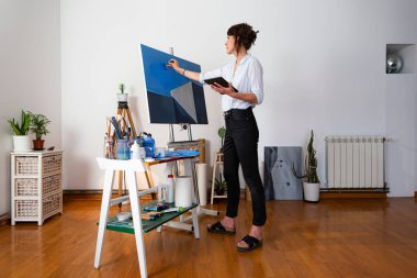 Genç ressam onun stüdyosunda duruyor ve tabletten eskizi doğrudan tablonun üzerine kopyalıyor. Arka planda resim yapmak için gerekli malzemeyi görebilirsiniz..