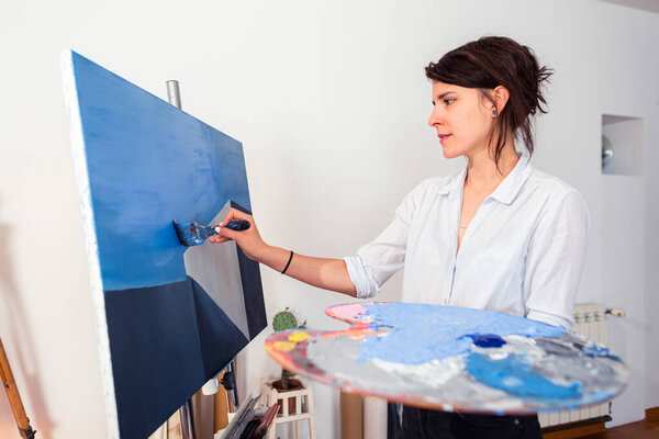 Молодая улыбающаяся художница наносит краску на холст в своей домашней студии. Вокруг него можно было увидеть и другие сопроводительные материалы для живописи. На некоторых фотографиях она держит палитру красок в одной руке, а с другой наносит краску с болью