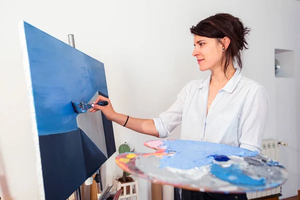 A young smiling painter applies paint to a canvas in her home studio. Other accompanying materials for painting could be seen around it. In some photos, she holds a palette of paints in one hand, while with the other she applies the paint with a pain
