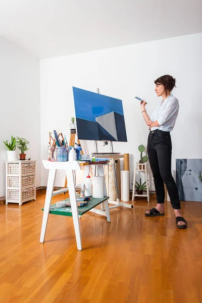 A young painter observes her work of art in her home studio. Around her, you can see a contemporary work of art placed on an easel, other pieces of art, as well as other accompanying material for painting.