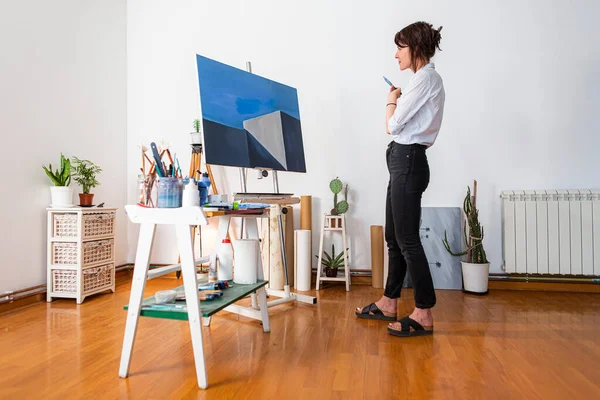 A young painter observes her work of art in her home studio. Around her, you can see a contemporary work of art placed on an easel, other pieces of art, as well as other accompanying material for painting.