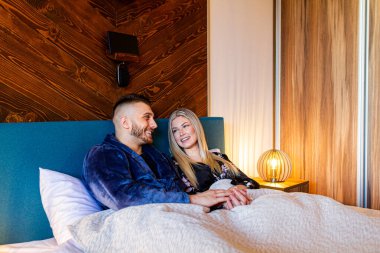 Yatakta yarı çıplak, beline kadar battaniyeyle örtülü genç, gülümseyen bir çift..