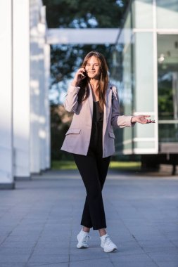 Genç ve güzel bir kız cep telefonuyla konuşurken yürüyor. Gülümsüyor, iş eğitimli, arka planda modern mimari var..
