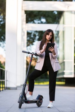 Genç ve güzel bir kız, bir ayağı ayakta duran bir platformda, elektrikli scooterının yanında duruyor. Gülümsüyor, iş eğitimli, arka planda modern mimari var..