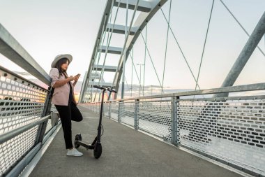 Genç bir kız köprü çitlerine yaslanıyor ve telefonunda daktilo yazıyor. Onun yanına elektrikli bir scooter park edilmiş. Nehir, günbatımı ve arka planda modern bir köprünün parçaları yer alıyor..