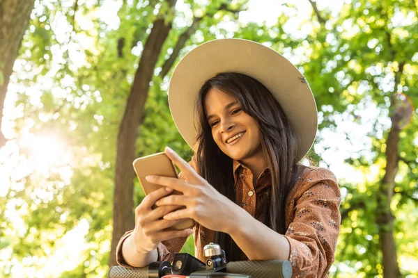 Telefonunda yazarken elektrikli scooterının yanında duran şapkalı güzel bir kız. Arka planda ağaçlar ve yeşil renkler baskın.