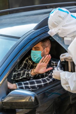 Adam sürüş için COVID 19 testi yapmayı reddediyor, hastadan araba camından burun örneği alıyor, PCR teşhisi koyuyor, PPE 'de elinde test kiti olan bir doktor.. 