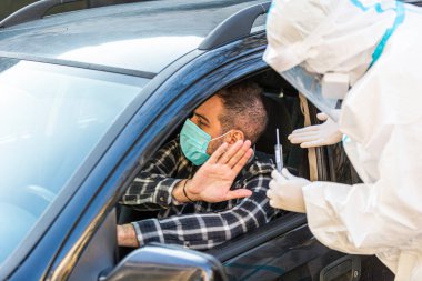 Adam sürüş için COVID 19 testi yapmayı reddediyor, hastadan araba camından burun örneği alıyor, PCR teşhisi koyuyor, PPE 'de elinde test kiti olan bir doktor.. 