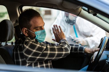 Adam sürüş için COVID 19 testi yapmayı reddediyor, hastadan araba camından burun örneği alıyor, PCR teşhisi koyuyor, PPE 'de elinde test kiti olan bir doktor..