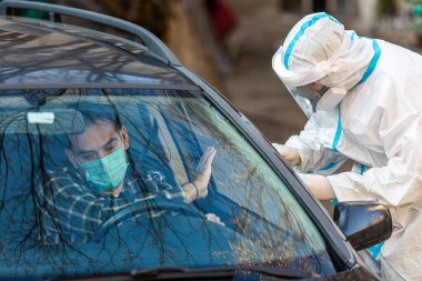 Adam sürüş için COVID 19 testi yapmayı reddediyor, hastadan araba camından burun örneği alıyor, PCR teşhisi koyuyor, PPE 'de elinde test kiti olan bir doktor..