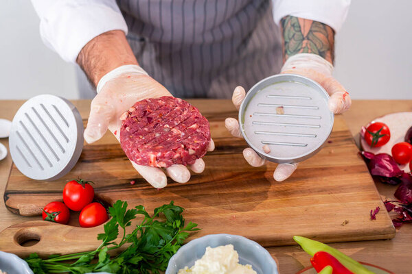 Close up of a photographed hand of a chef showing a ready raw burger just taken out of a shaping press. Vegetables and primrose leaves can be seen around the kitchen wooden board.