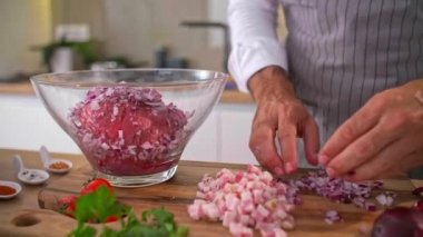 Aşçı soğanı şeffaf cam bir kaseye koyuyor, iş yüzeyinde sebzeler, pastırma dilimleri, baharatlar ve küçük mutfak aletleri var..