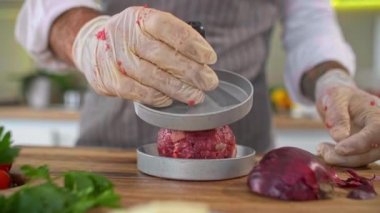 Mutfaktaki ahşap bir tahtanın üzerinde hamburgerler için et şekillendiren bir şefin yakın çekimi. Çeşitli baharatlar, sebzeler ve iş yüzeyinin etrafında küçük mutfak kaseleri var..