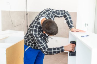 Marangoz vidaları vidalıyor ve mutfak parçalarını elektrikli tornavidayla birleştiriyor. Etrafında iş aletleri ve çalıştığı malzemeler var..