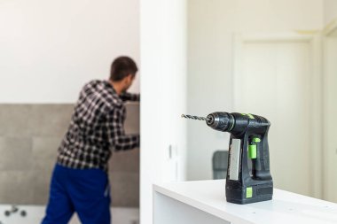 Matkap dairenin odasındaki çalışma yüzeyine yerleştirilir, arka planda ise rafları yerleştirmek için duvarı delme işini yapan bir marangoz görebilirsiniz..
