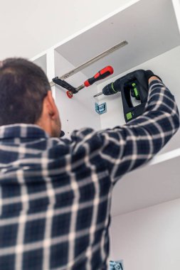 Marangoz vidaları vidalıyor ve mutfak parçalarını elektrikli tornavidayla birleştiriyor. Etrafında iş aletleri ve çalıştığı malzemeler var..