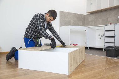 Marangoz, yeni bitmiş mutfak için rafları ve dolapları monte etmeye başladığı boş bir dairede rafları ölçüyordu. Etrafında iş aletleri var..