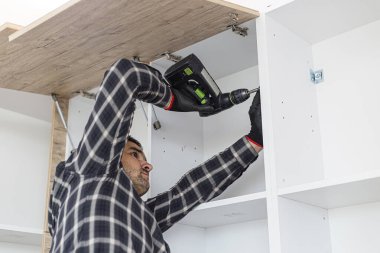 Marangoz vidaları vidalıyor ve mutfak parçalarını elektrikli tornavidayla birleştiriyor. Etrafında iş aletleri ve çalıştığı malzemeler var..