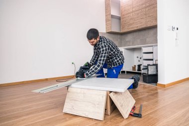 Bir marangoz odanın zemininde çalışma yüzeyinin ahşap bir tahtasını kesiyor. İşini, kendisine bir çalışma masası olarak hizmet eden ahşap bir kutuda yapar..