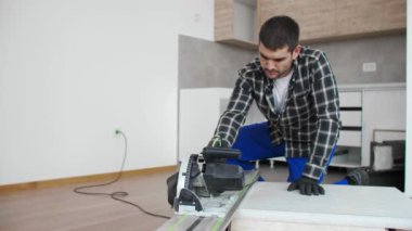 Bir marangoz odanın zemininde çalışma yüzeyinin ahşap bir tahtasını kesiyor. İşini, kendisine bir çalışma masası olarak hizmet eden ahşap bir kutuda yapar..