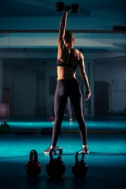 Genç bir bayan gülümsüyor ve spor salonunda bir aynanın önünde halterle egzersiz yapıyor..