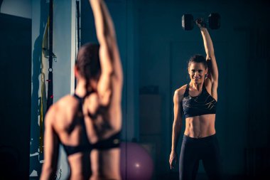 Genç bir bayan gülümsüyor ve spor salonunda bir aynanın önünde halterle egzersiz yapıyor..