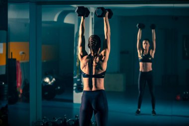 Spor salonundaki aynanın önünde iki dambılla egzersiz yapan genç bir kadın..
