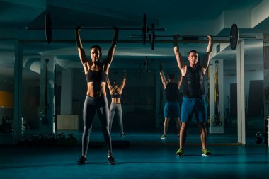 Spor salonunda antrenman yapan bir çift halterin ağırlığıyla üstten çömelme egzersizi yapıyor..