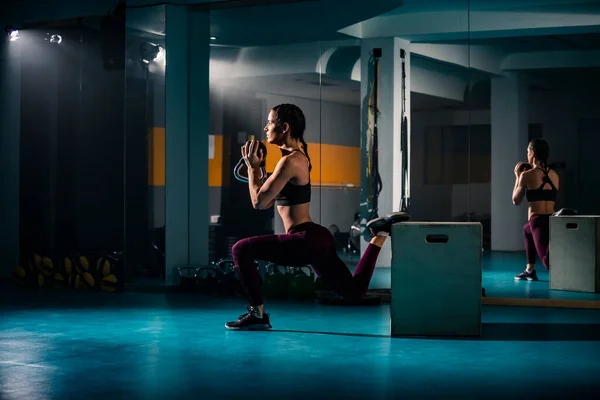 Etrafında ağırlık ve diğer egzersiz ekipmanlarıyla boş spor salonunda çömelme egzersizleri yapan genç bir kadın..