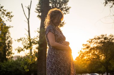 Gün batımında arka planda ağaçlarla birlikte şehir parkında duran mutlu, beyaz hamile bir kadın. Gülümsüyor, aşağı bakıyor ve kollarını karnına sarıyor..