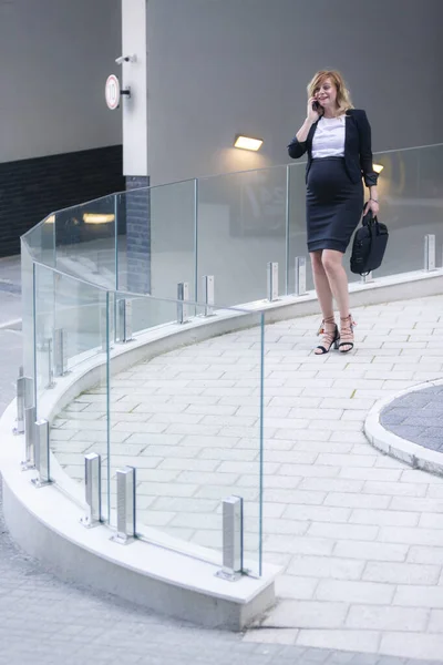 A smiling pregnant businesswoman walking in front of the modern building and talking on her mobile phone.